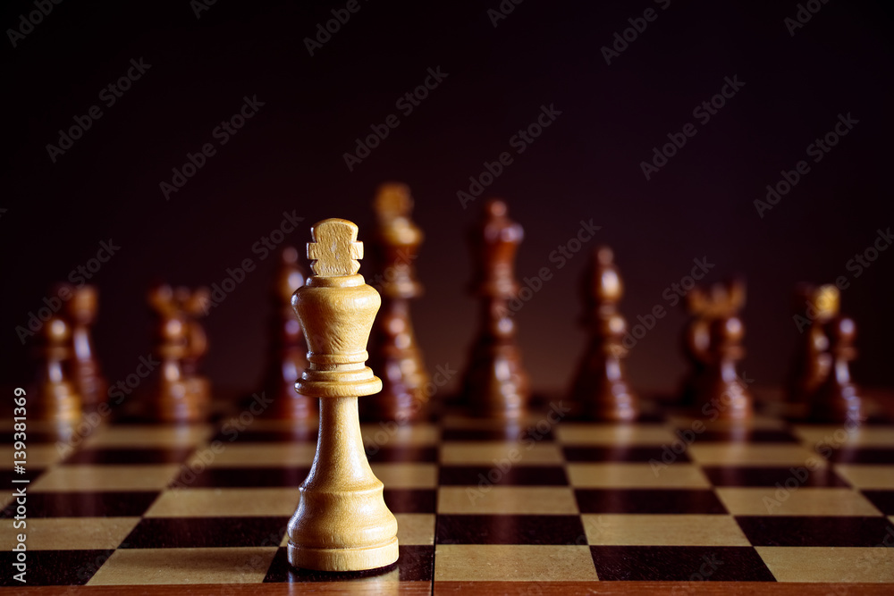 White chess king in front of a chessboard, black chess pieces on a dark background White chess king in front of a chessboard, black chess pieces on a dark background