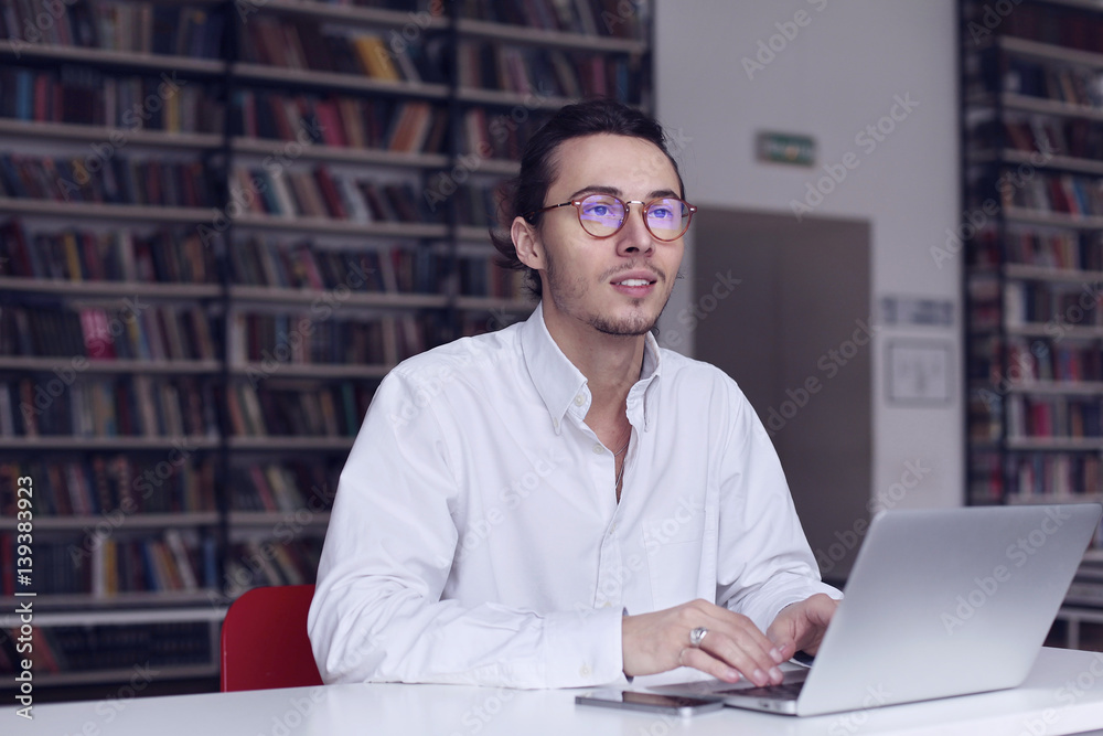 Young entrepreneur, university student working on laptop with book on the scientific thesis in a library Young entrepreneur, university student working on laptop with book on the scientific thesis in a library