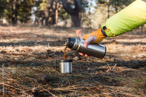 A thermos and a cup with hot coffee in the forest background.