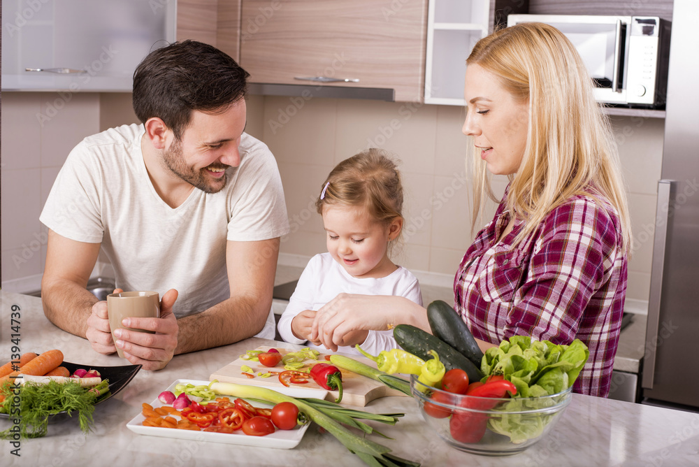 Young couple with kid having fun in kitchen while preparing fresh vegetable salad