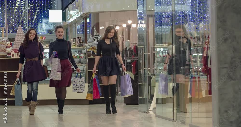 Happy female shoppers walk at shopping mall with sale bags HD video. Young girl friends talk, smile and enjoy purchases going past boutique showcase