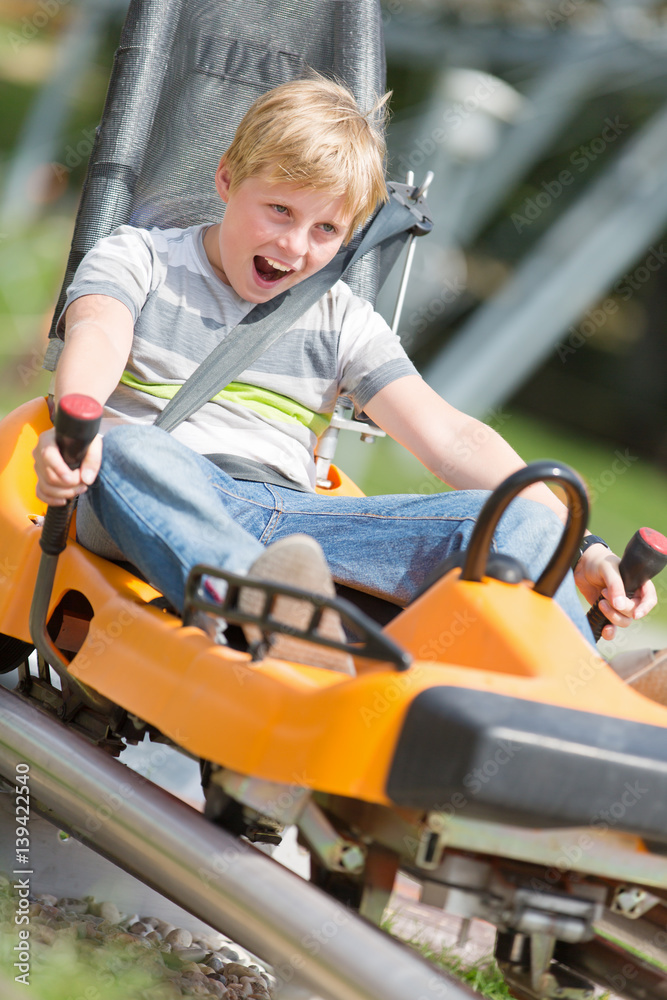 Happy Boy Riding at Bobsled Roller Coaster Rail Track in Summer Amusement Park Happy Boy Riding at Bobsled Roller Coaster Rail Track in Summer Amusement Park