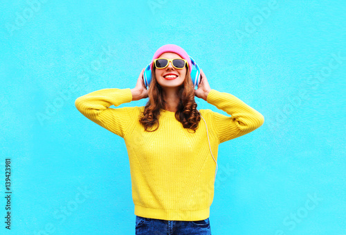 Fashion smiling woman listening to music in headphones wearing a colorful pink hat, yellow sunglasses and sweater over blue background