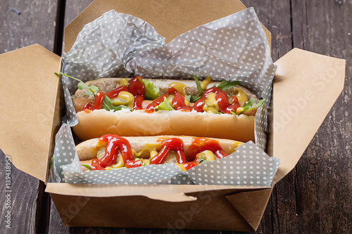 Home Made Hot Dogs with tomatoes, chili gherkins wrapped in the paper and in the box ove a rustic wooden background