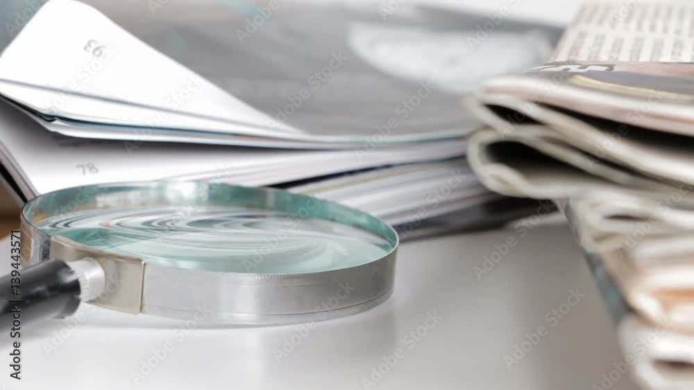Magnifier - reading glass - and stacked newspapers on white table. Selective/switched focus. Magnifier - reading glass - and stacked newspapers on white table. Selective/switched focus.