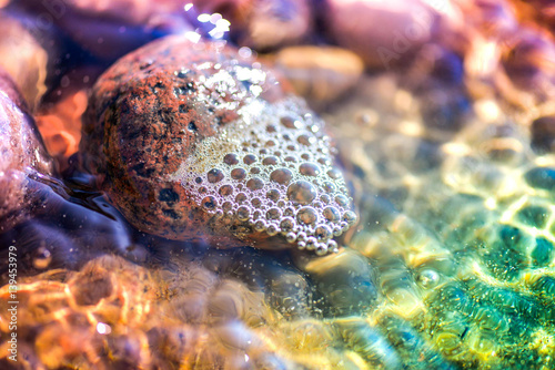 Macro shot of magical, healing sea pebbles and rocks, white bubbles and foam in colorful, vibrant ocean water full of energy. Sun rays, reflections deep in the salty water at the beach during sunset