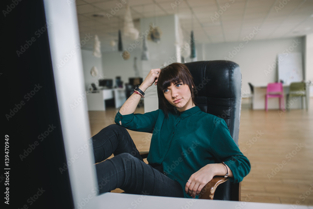 The young beautiful girl worker in a dark jacket sitting at work in an office chair and looking at the big monitor. The modern office worker. The young beautiful girl worker in a dark jacket sitting at work in an office chair and looking at the big monitor. The modern office worker.