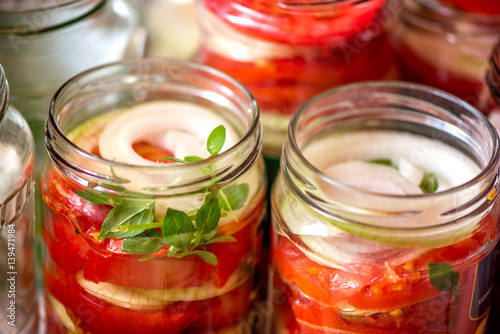 Canning fresh tomatoes with onions for winter in jelly marinade. A shot of basil leaves on top of a red ripe tomato slices and onion rings being put in jar.