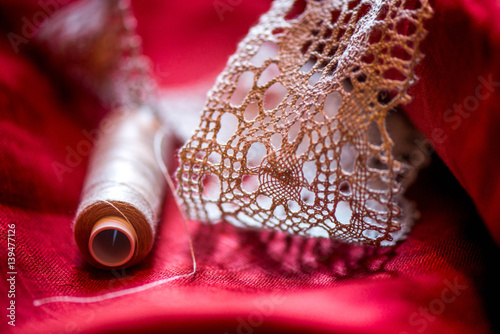 A macro shot of a white thread spool and a white, crochet natural linen lace trim on a dark red linen fabric, being used while sewing a pillowcase by using a sewing machine