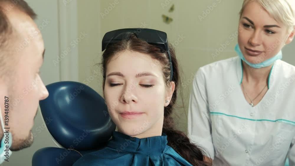 Patient communicate with her dentist in dental office. Portrait. Brunette adult lady in green silk blouse with braided hair sitting in special chair talk to doctor, handsome man with Spanish mustache Patient communicate with her dentist in dental office. Portrait. Brunette adult lady in green silk blouse with braided hair sitting in special chair talk to doctor, handsome man with Spanish mustache