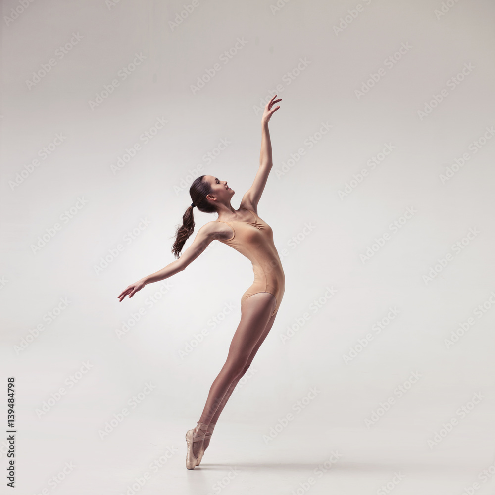 young beautiful ballet dancer in beige swimsuit posing on pointes on light grey studio background