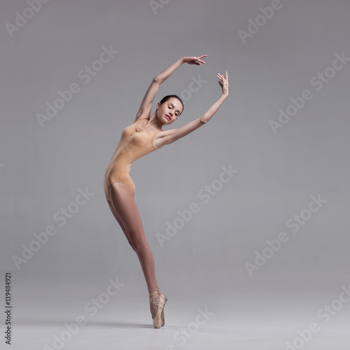 young beautiful ballet dancer in beige swimsuit posing on pointes on light grey studio background