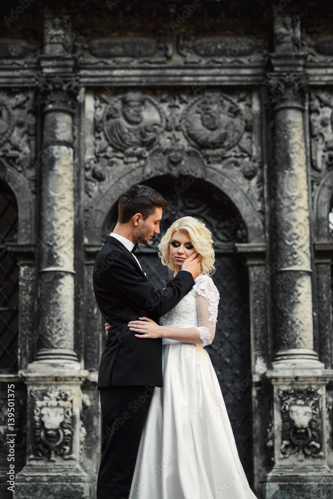 Handsome groom holds charming bride's head tenderly while standing before historic building
