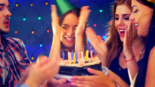 Happy friends birthday party with candle celebration cakes. Girl girl joyfully clapping. People looking at burning candles in night spot. Two women and man have fun.