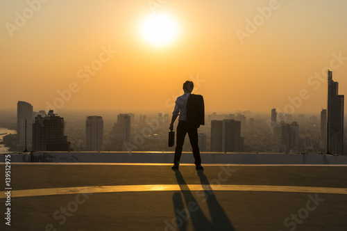 Silhouette Rear view of businessman standing with carrying the business bag and looking the vision over the cityscape background at sunset time with lens flare,Business success concept