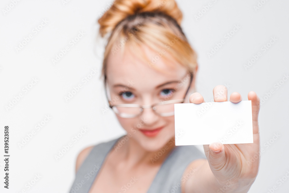 Nice young girl in glasses and vest showing white empty card with copy space. Blurred portrait on grey background, focus on blank paper, face close up. Nice young girl in glasses and vest showing white empty card with copy space. Blurred portrait on grey background, focus on blank paper, face close up.