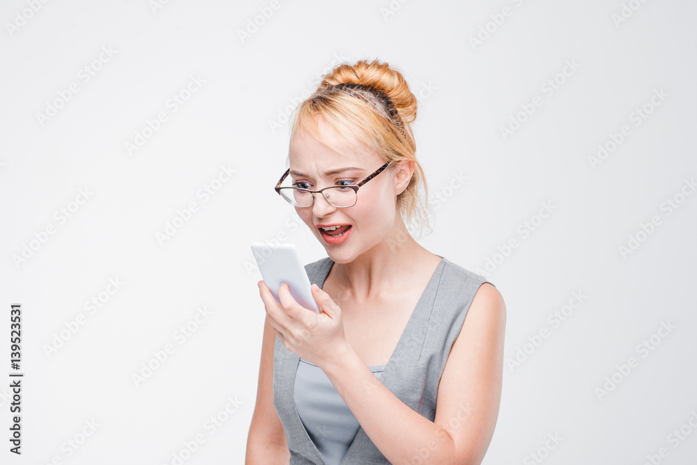 Irritated young girl in glasses with mobile phone looks angry, peeved and irritated. Annoyed and grumpy blonde with smartphone on grey background with copy space. Irritated young girl in glasses with mobile phone looks angry, peeved and irritated. Annoyed and grumpy blonde with smartphone on grey background with copy space.