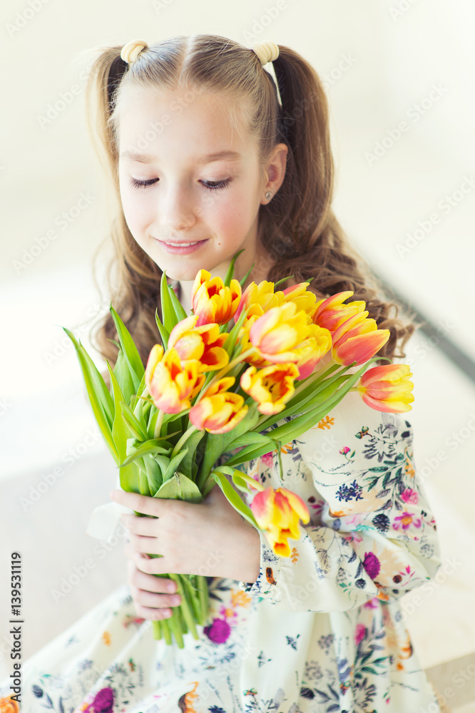 Beautiful toddler girl sitting in the room with fresh tulips, smiling with happy face, waiting for spring and Easter holidays Beautiful toddler girl sitting in the room with fresh tulips, smiling with happy face, waiting for spring and Easter holidays