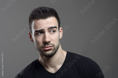 Moody portrait of confused man facial reaction looking away over dark gray studio background.