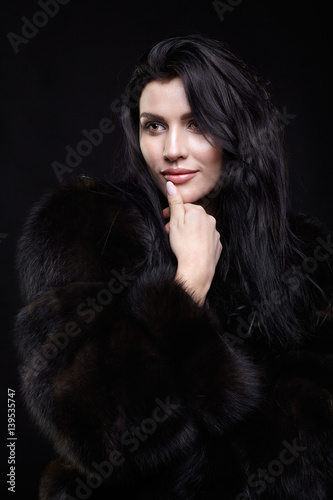 Portrait of a young brunette woman with long black hair dressed in a fur coat on black