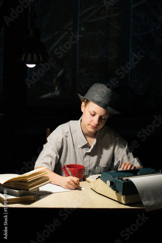 Portrait of a girl in a hat sitting at a table with a typewriter and books, making notes at night