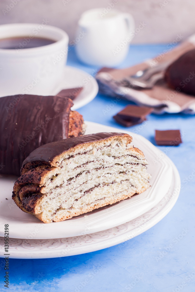 Homemade pastry with poppy seeds for breakfast icing chocolate on white plate with cup of coffee and milk on blue table background. copy space. Homemade pastry with poppy seeds for breakfast icing chocolate on white plate with cup of coffee and milk on blue table background. copy space.