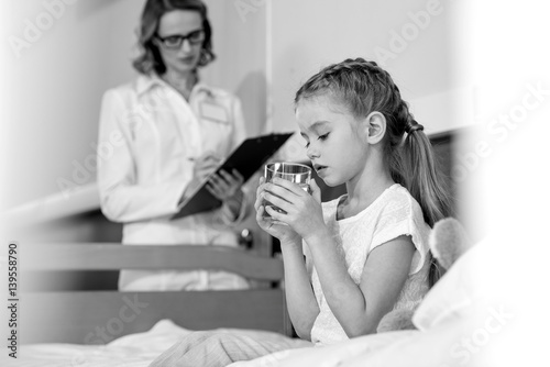 Doctor writing in clipboard while little girl patient taking medicines, black and white photo