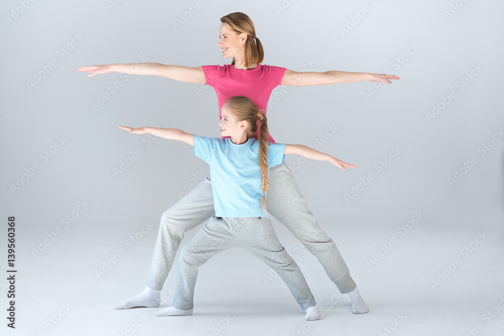 Fototapeta premium Side view of sporty mother and daughter practicing warrior yoga pose together on white