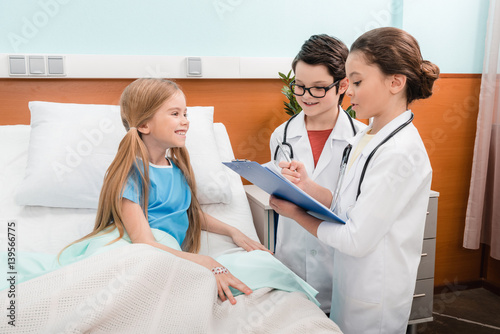 Boy and girl doctors writing in clipboard while standing near smiling little patient
