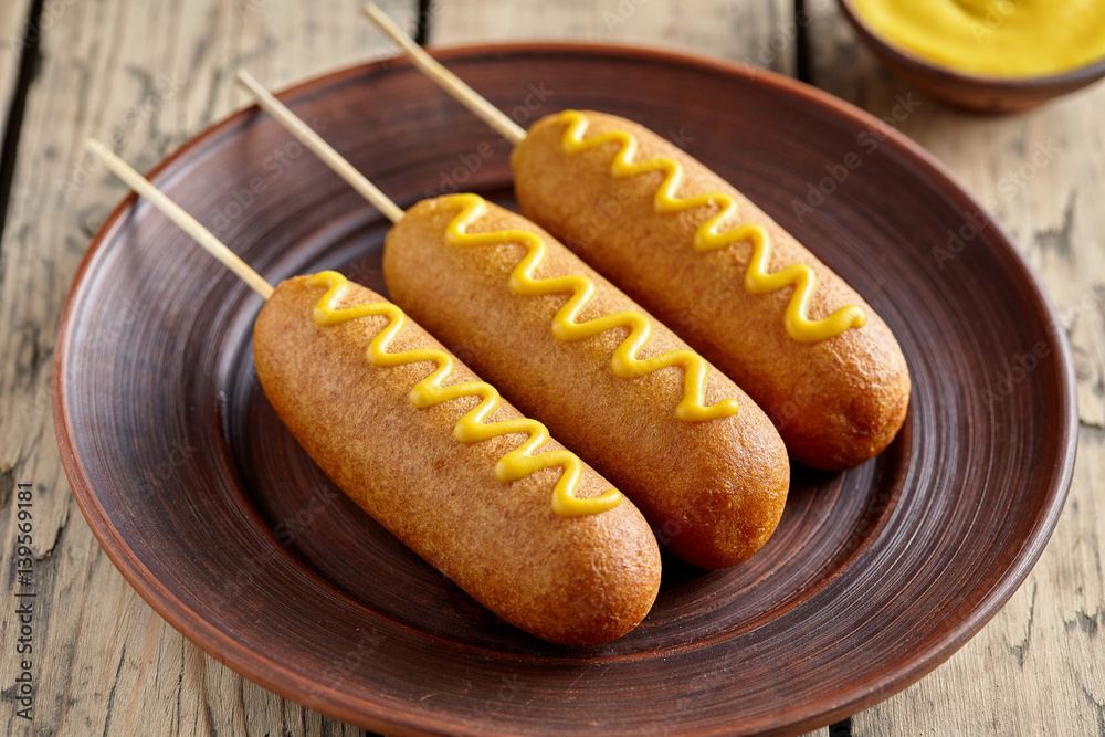 Corn dog traditional American corndog street junk food deep fried hotdog meat sausage with mustard snack treat coated in layer of cornmeal batter on stick unhealthy eating on rustic wooden desk. Corn dog traditional American corndog street junk food deep fried hotdog meat sausage with mustard snack treat coated in layer of cornmeal batter on stick unhealthy eating on rustic wooden desk.