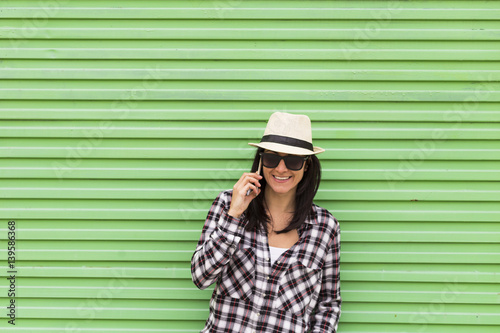 Happy beautiful woman talking on the phone over green background. Wearing hat and sunglasses.