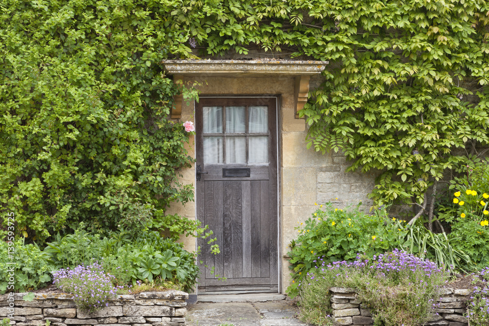 Obraz premium Old brown wooden doors in English stone house with flowers in the front garden, surrounded by green climber .