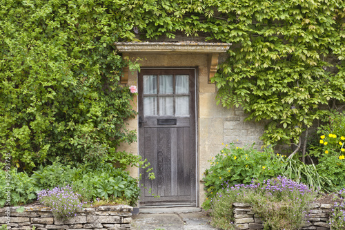 Old brown wooden doors in English stone house with flowers in the front garden, surrounded by green climber .
