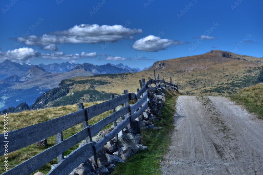 Straße mit einem Zaun in Richtung Berge gegen den blauen Himmel