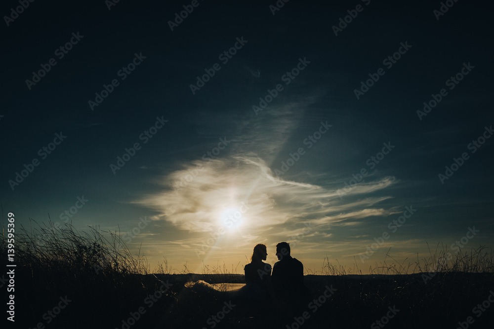 groom and bride in a wedding dress sitting in a field at sunset