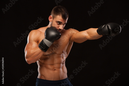 Muscular young man with perfect Torso with six pack abs, in boxing gloves is showing the different movements and strikes isolated on black background with copyspace