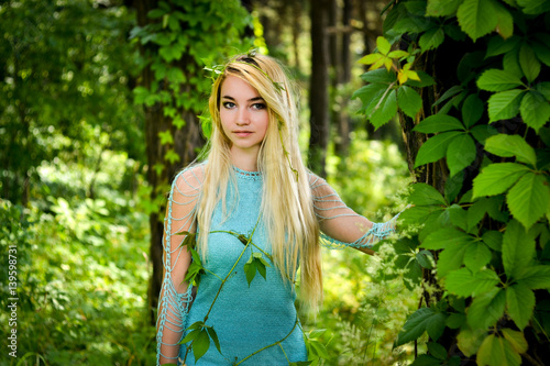 Pretty young blonde girl with long hair in turquoise dress standing in the green forest where trees are enlaced with lianas photo