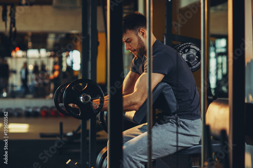 Handsome man doing biceps lifting barbell on bench in a gym