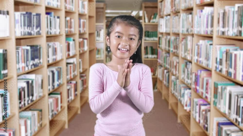 Happy female elementary school student standing in the library while clapping hands Happy female elementary school student standing in the library while clapping hands
