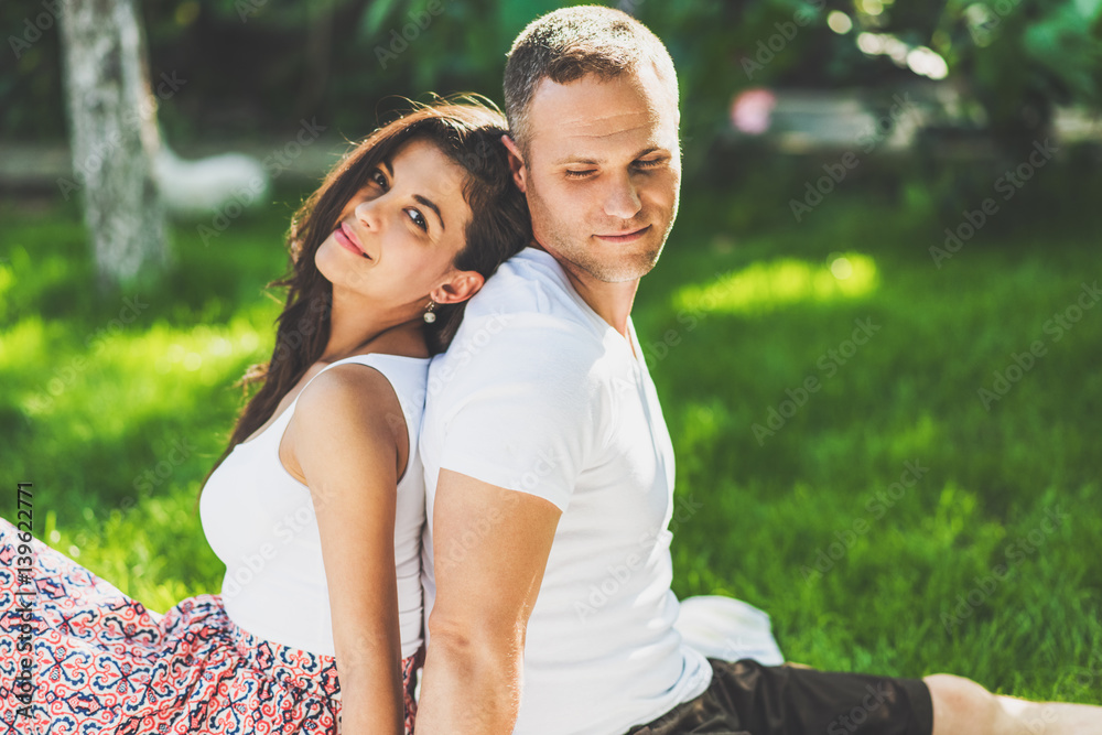 Cute couple in love sitting on a picnic blanket outdoor. Caucasian happy dreamy man and smiling brunette woman sitting on green grass in park. Cute couple in love sitting on a picnic blanket outdoor. Caucasian happy dreamy man and smiling brunette woman sitting on green grass in park.