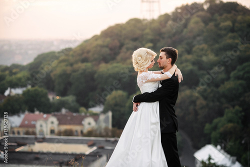 Fototapeta Sensual beautiful bride and handsome groom hugging tenderly on a hilltop overloo