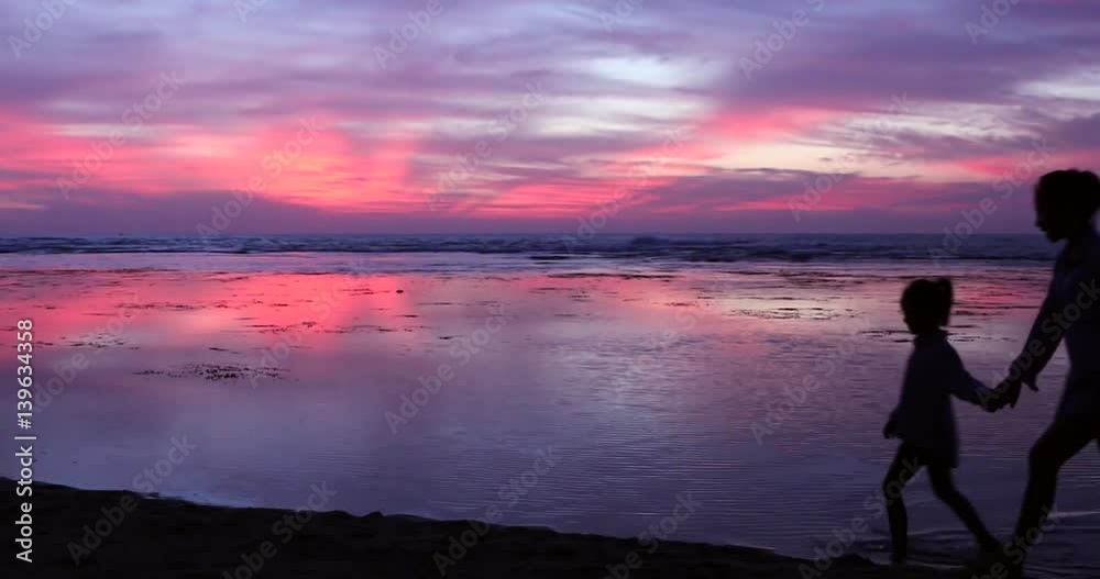 Silhouette of young woman and her daughter walking on the beach while holding hands with beautiful sky at dusk time Silhouette of young woman and her daughter walking on the beach while holding hands with beautiful sky at dusk time