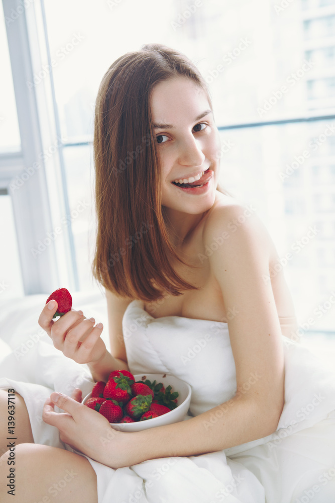 Portrait of smiling beautiful young Caucasian woman with long hair sitting in bed early morning by window, eating red fresh strawberry, showing her tongue flirting, lifestyle, toned with filters