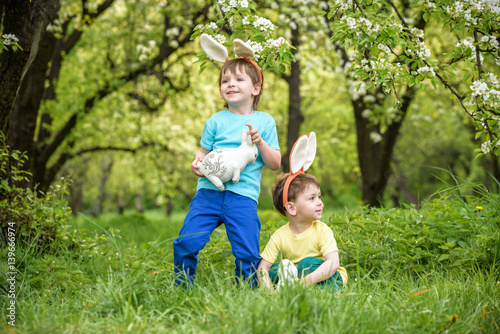 Two little kids boys and friends in Easter bunny ears during traditional egg hunt in spring garden, outdoors. Siblings having fun with finding colorful eggs. Old christian catholoc tradition