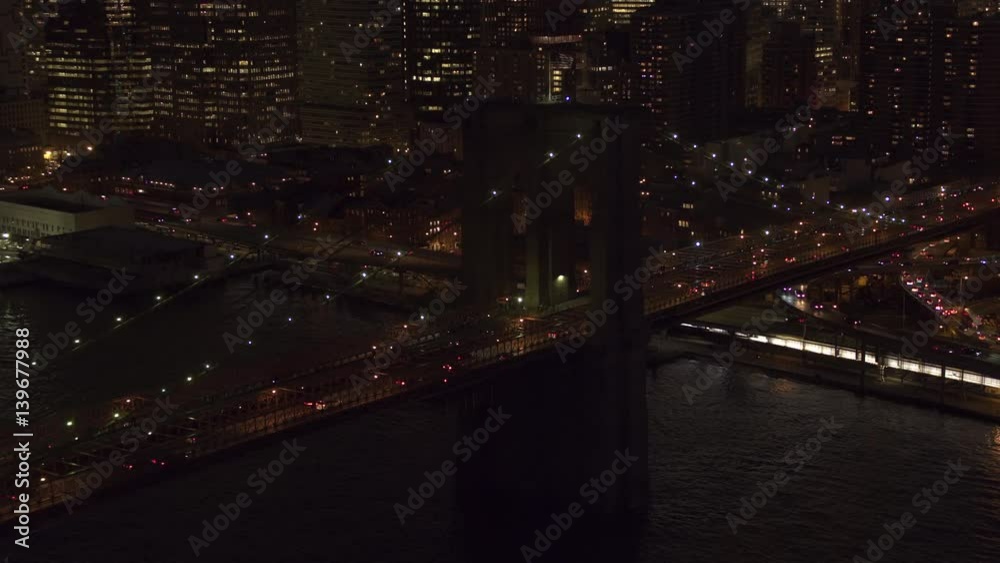 AERIAL HELI SHOT, CLOSE UP: Flying above busy jammed interstate on iconic Brooklyn Bridge lit up with lights leading to Downtown Manhattan business district. Commuters traveling to work before sunrise