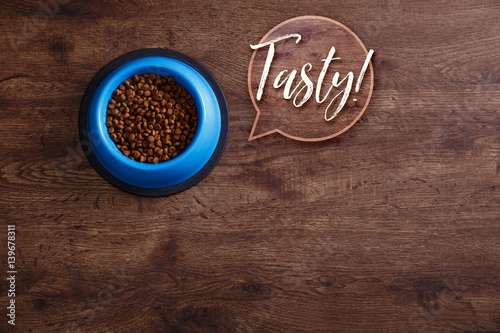 Bowl of dry kibble dog food. Tasty speech bubble. Healthy pets feed. Blue plate on wooden rustic background.