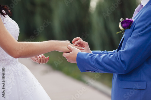 Wedding rings. The groom in a suit put the ring to the bride in a white dress standing and holding hands in the green garden.
