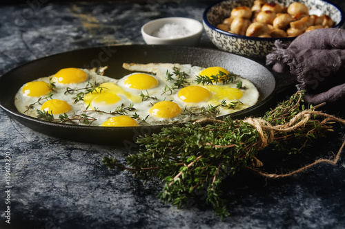 Fried quail eggs in a frying pan with thyme and mushrooms. Dark background.