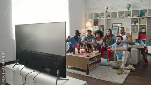 Multi-ethnic group of young flat mates watching sports competition on TV at home. They jumping up, raising hands and yelling while celebrating goal of favorite team