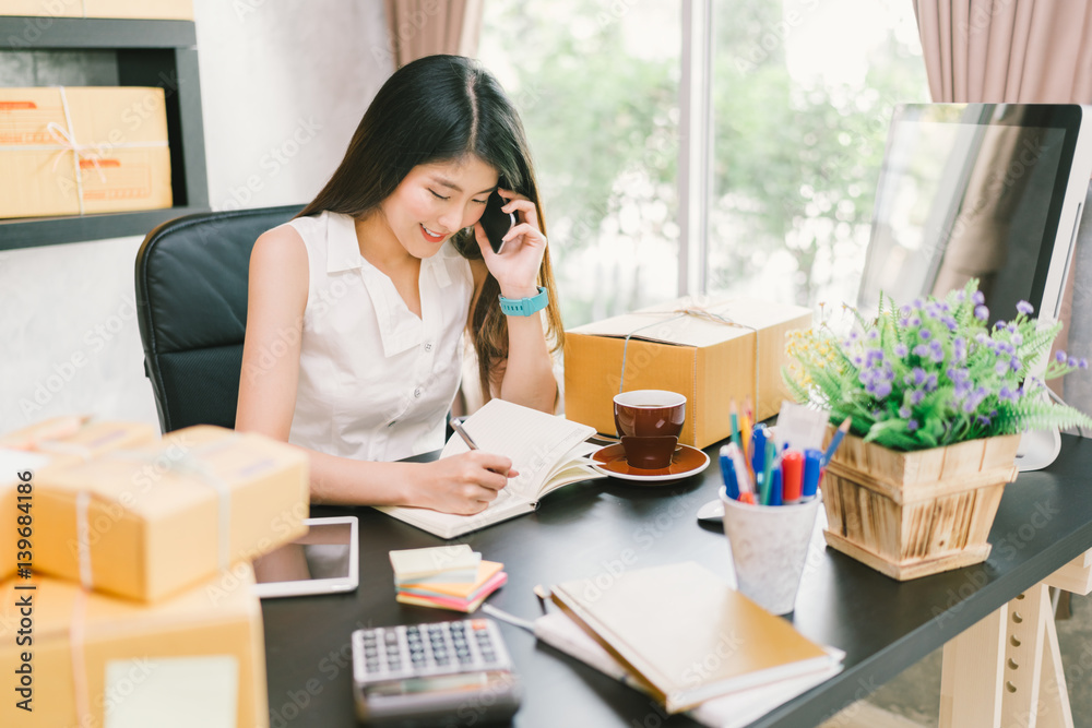 Young Asian small business owner working at home office, using mobile phone and taking note on purchase orders. Online marketing packaging delivery, startup SME entrepreneur or freelance woman concept Young Asian small business owner working at home office, using mobile phone and taking note on purchase orders. Online marketing packaging delivery, startup SME entrepreneur or freelance woman concept